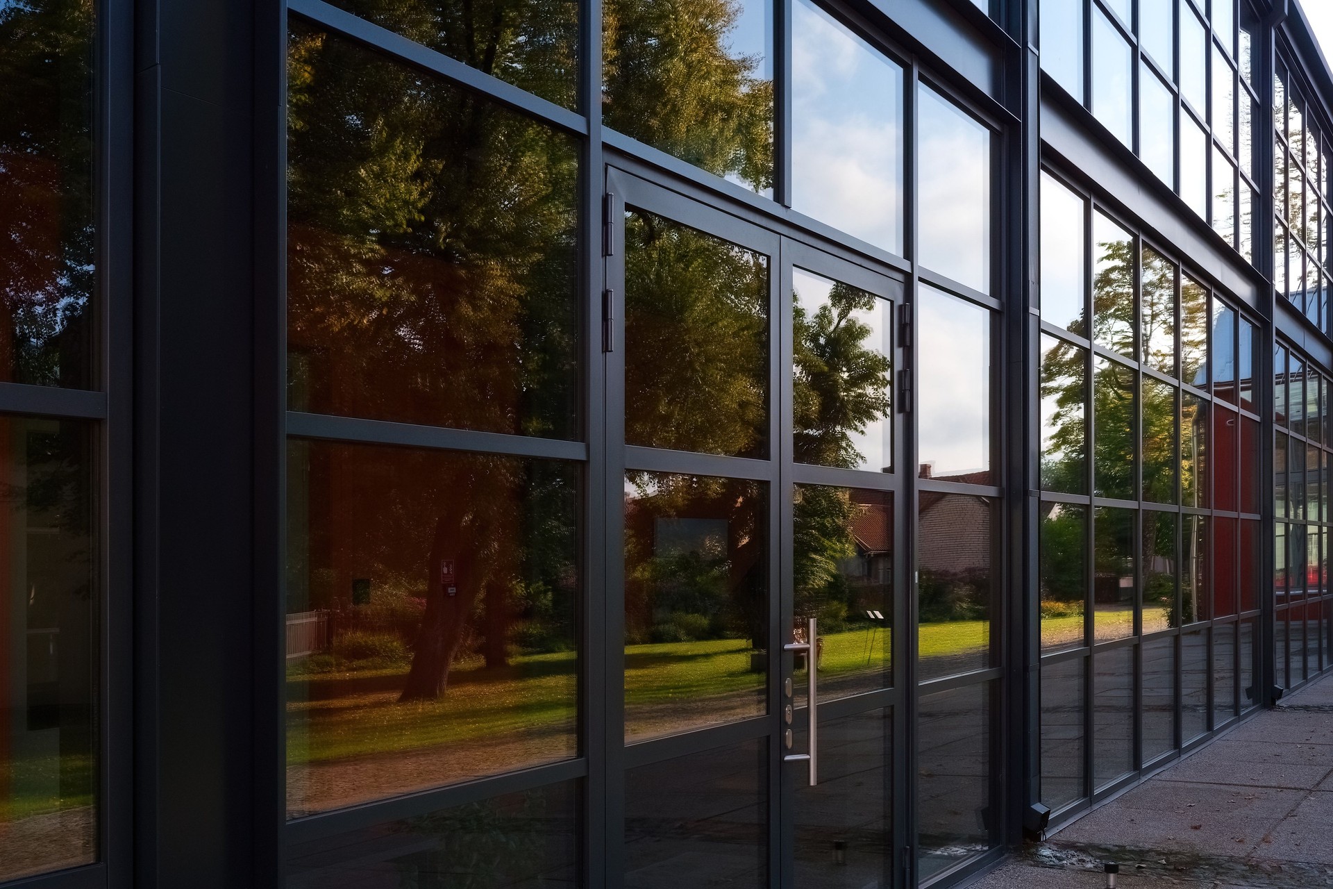 The modern glass facade of a building with a black metal frame reflects the surrounding trees and a small house. The perspective view showcases the clean lines and contemporary architectural design.